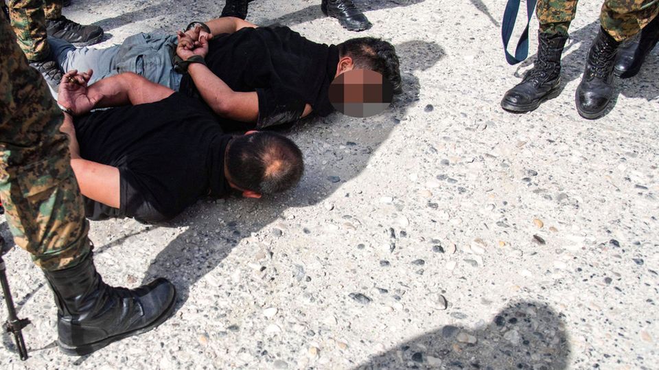 Soldiers in camouflage and with black stiffeners stand around two tied men lying on the ground
