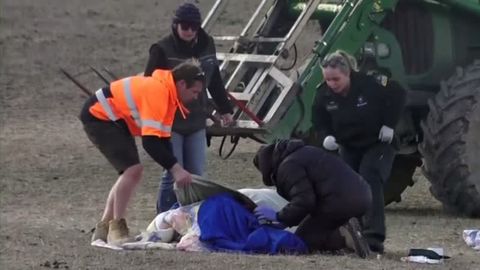 Zwei Männer und zwei Frauen beugen sich über eine blau gekleidete, am Boden liegende Person. Im Hintergrund steht ein Traktor.