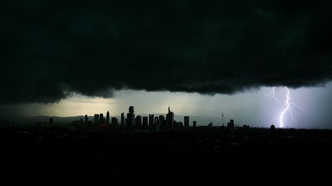Tiefschwarze Gewitterwolken über einem Stadtpanorama, rechts ist ein Blitz zu sehen