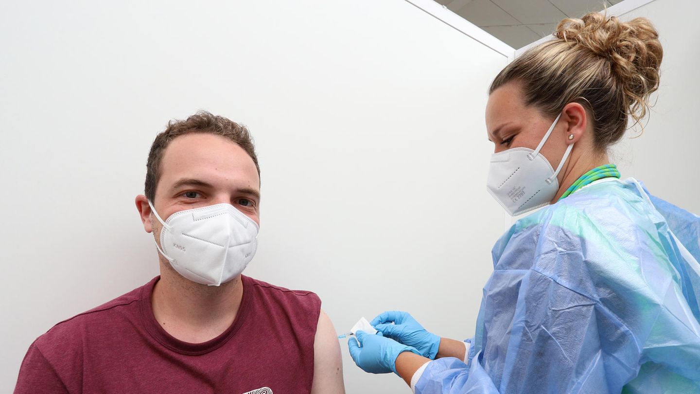 Ein junger weißer Mann mit FFP2-Maske und dunkelrotem T-Shirt, während eine blonde Frau ihm eine Spritze in den Oberarm sticht