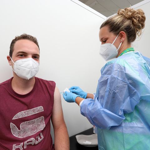 Ein junger weißer Mann mit FFP2-Maske und dunkelrotem T-Shirt, während eine blonde Frau ihm eine Spritze in den Oberarm sticht