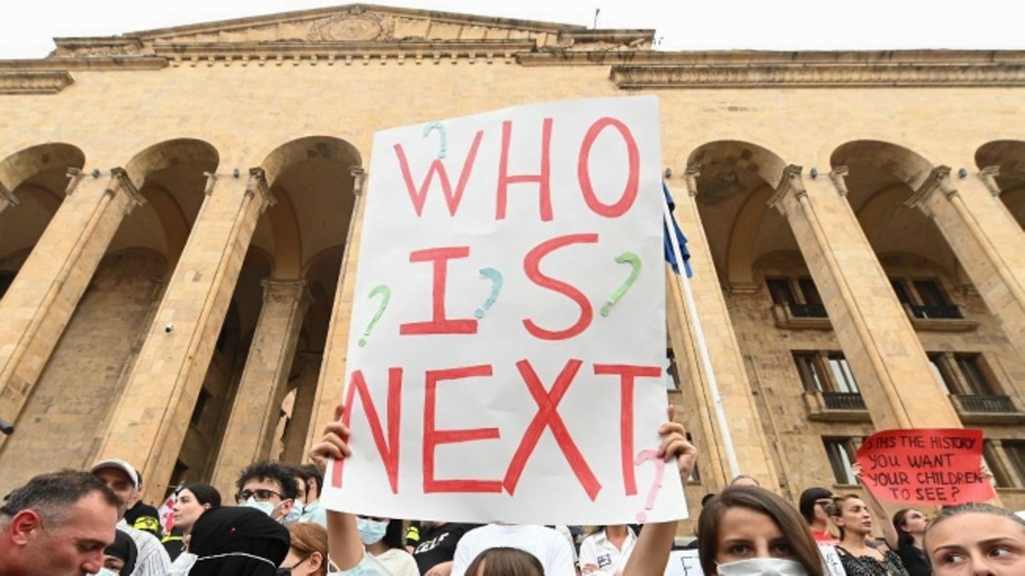Mehrere Menschen stehen vor einem Gebäude und demonstrieren, eine Frau hält ein Schild, auf dem "who is next?" steht