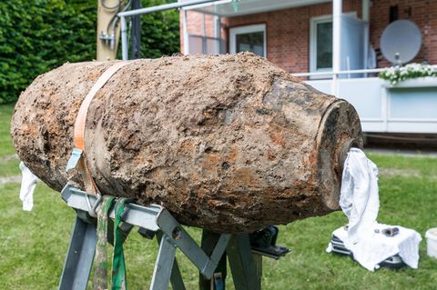 Eine mit Erde und Rost überzogene Bombe liegt auf einem Gestell vor einem Mehrfamilienhaus