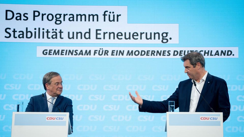 Auf einer Bühne mit hellblauem Hintergrund stehen zwei Rednerpulter, an denen zwei weiße Männer in Anzügen stehen