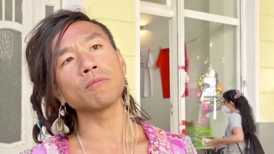 Viet Dinh Khac Ein asiatisch aussehender Mann mit längeren Haaren und Feder-Ohrring legt den Kopf schräg und steht er vor einem gelben Altbau