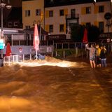 Drei Personen machen Fotos und beobachten die Wassermassen, die sich über eine Straße und ein Restaurant ausbreiten