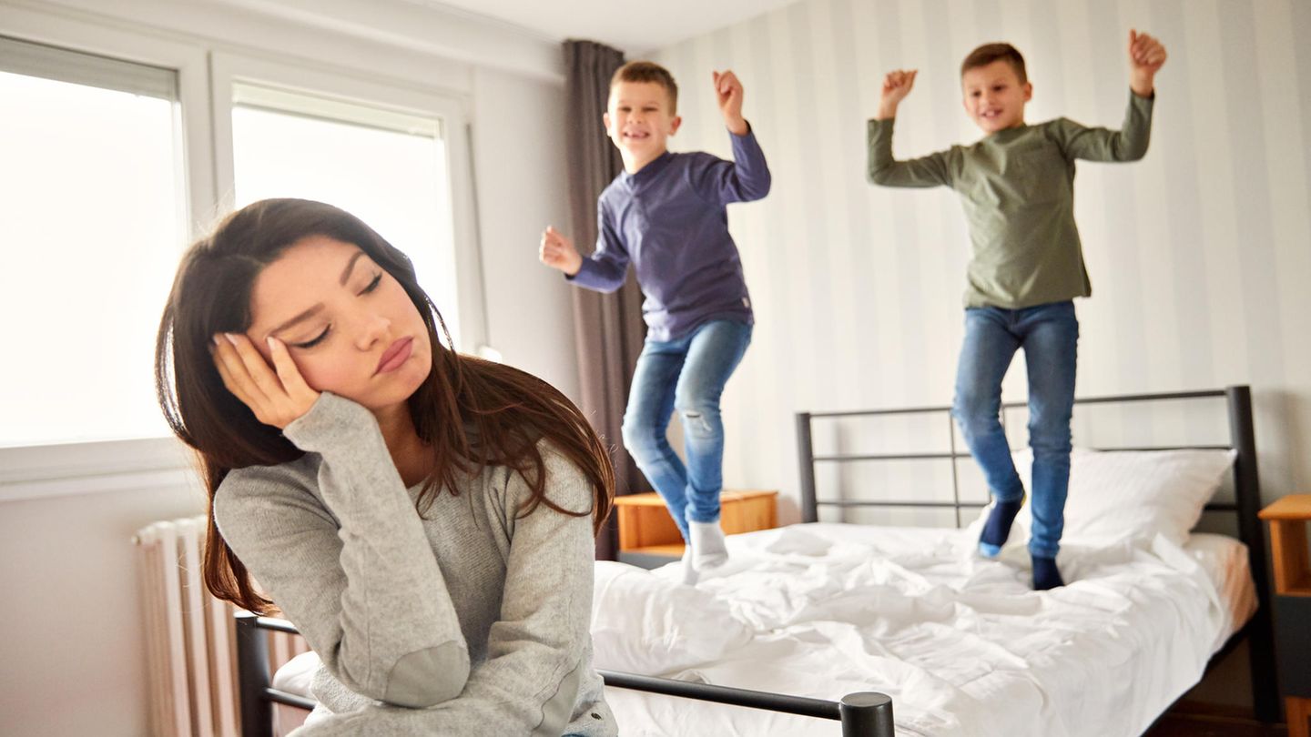 Mutter sitzt müde vor dem Bett ihrer Kinder, die im Hintergrund darauf herumspringen.