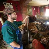 Ein junger Mann mit Vollbart und Cap steht auf eine Schuafel gestützt in einem von Wasser verwüsteten Wohnzimmer