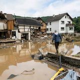 Ein Mann in Gummistiefeln und Arbeitshose geht auf einem Mäuerchen auf ein vom Wasser zerstörtes Fachwerkhaus zu