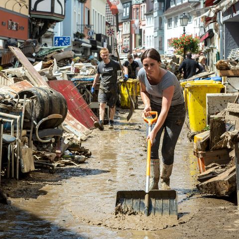 Mit einem Schneeschieber reinigt eine weiße Frau in Gummistiefeln eine Straße von schlammigem Wasser - oder versucht es