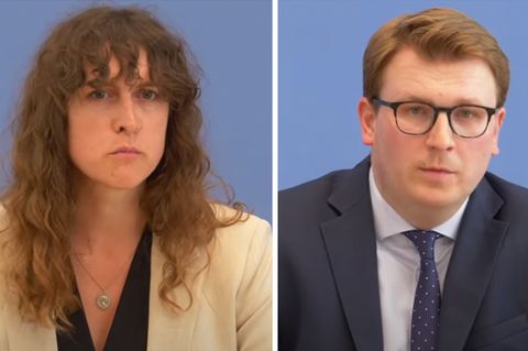 Eine weiße Frau mit halblangen, braunen Locken und ein weißer Mann mit schwarzer Brille und blondem Seitenscheitel sitzen