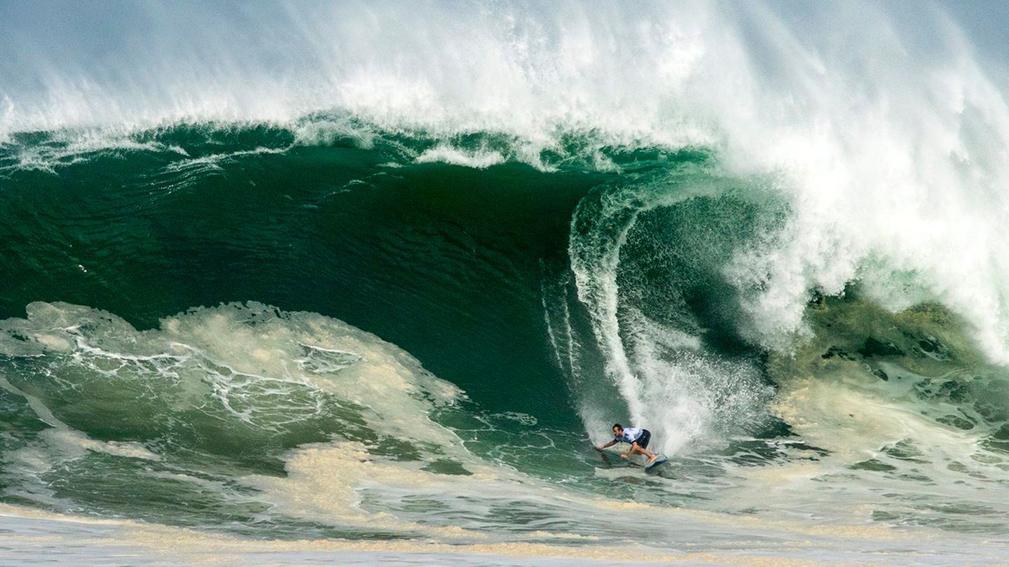 Ein Surfer erscheint sehr klein, während er eine Riesenwelle hinabsurft, von deren Kamm Gischt abweht