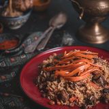 Qabuli Palau (Reis mit Lamm, Karotten und Rosinen)      Zutaten:   500 g Sella-Reis (im afghanischen Supermarkt oder online erhältlich), 1 kg Lammfleisch aus der Keule mit Knochen (vom Metzger in 5 cm große Würfel schneiden lassen), 3 Zwiebeln, Pflanzenöl zum Anbraten, 2–3 TL Char Masala oder Garam Masala, 1 TL Kardamompulver, frisch gemahlener schwarzer Pfeffer, Salz, 200 g Rosinen, 4 Bio-Karotten (insgesamt 400 g), 4 TL Zucker      Zubereitung:   1. Den Reis mit Wasser bedecken und 1–2 Stunden quellen lassen. Anschließend abgießen und abtropfen lassen.  2. Das Lammfleisch trockentupfen. Die Zwiebeln abziehen und in feine Würfel schneiden. Etwas Öl in einem Schnellkochtopf erhitzen und das Fleisch darin rundherum kräftig anbraten. Dann die Zwiebeln dazugeben und langsam goldbraun schmoren. Die Mischung mit dem Char oder Garam Masala, dem Kardamompulver, Pfeffer und Salz würzen. 750 ml Wasser angießen und das Lamm im fest verschlossenen Schnellkochtopf 20 Minuten garen. (Bei Zubereitung in einem normalen Topf beträgt die Garzeit 60–90 Minuten.)  3. In der Zwischenzeit bei Bedarf die Rosinen entstielen. Die Karotten waschen, putzen und zunächst längs in dünne Scheiben und dann in schmale Streifen schneiden. Etwas Öl in einer Pfanne erhitzen. Die Rosinen darin unter ständigem Rühren so lange braten, bis sie prall und rund geworden sind. Anschließend herausnehmen und beiseitestellen. Die Karotten in derselben Pfanne etwa 3–4 Minuten anbraten. Zuletzt den Zucker unterrühren und die Karotten 2 Minuten karamellisieren. Anschließend herausnehmen und zusammen mit den Rosinen bis zum Anrichten beiseitestellen.  4. Nach Ende der Garzeit des Lamms am Schnellkochtopf den Druck ablassen. Den Schnellkochtopf öffnen, das Fleisch aus dem Sud heben und abgedeckt warm halten. Den Sud durch ein feines Sieb in einen großen Topf streichen und kurz aufkochen. Dann den Reis zum Sud geben. Bei aufgesetztem Deckel bei mittlerer Temperatur so lange garen, bis die Flüssigkeit vom Reis komplett aufgenommen ist.  5. Das Fleisch auf den Reis setzen. Den Topfdeckel mit einem Küchentuch umwickeln und auf den Topf setzen. Alles bei sehr niedriger Temperatur weitere 10–15 Minuten garen.  6. Zum Anrichten die Fleischstücke auf einen großen Teller geben und mit dem Reis bedecken. Den Qabuli Palau mit den Rosinen und den karamellisierten Karottenstreifen bestreuen und servieren.