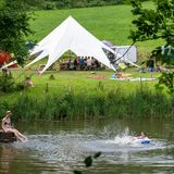Bild 1 von 9 der Fotostrecke zum Klicken:  Teilnehmer eines Ferienlagers in Rosenberg in Baden-Württemberg schwimmen in einem See, während die anderen zusammen im Gemeinschaftszelt sitzen.