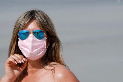 Eine weiße Frau mit langen, dunkelblonden Haaren trägt am Strand eine verspiegelte Piloten-Sonnenbrille und eine OP-Maske