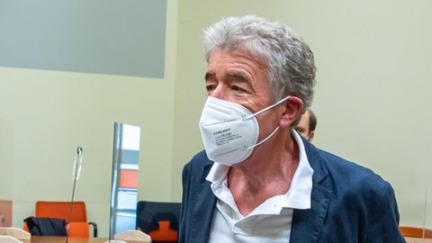 Ein älterer Mann mit weißem Bürstenschnitt und blauem Anzug steht mit FFP2-Maske in einem Gerichtssaal