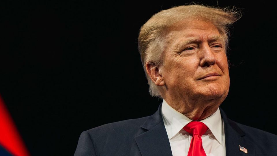 Milde lächelnd steht Donald Trump in Anzug und mit typischer roter Krawatte an einem Rednerpult vor dunklem Hintergrund