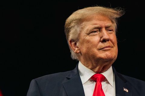Milde lächelnd steht Donald Trump in Anzug und mit typischer roter Krawatte an einem Rednerpult vor dunklem Hintergrund
