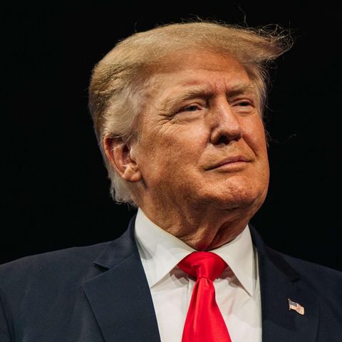 Milde lächelnd steht Donald Trump in Anzug und mit typischer roter Krawatte an einem Rednerpult vor dunklem Hintergrund