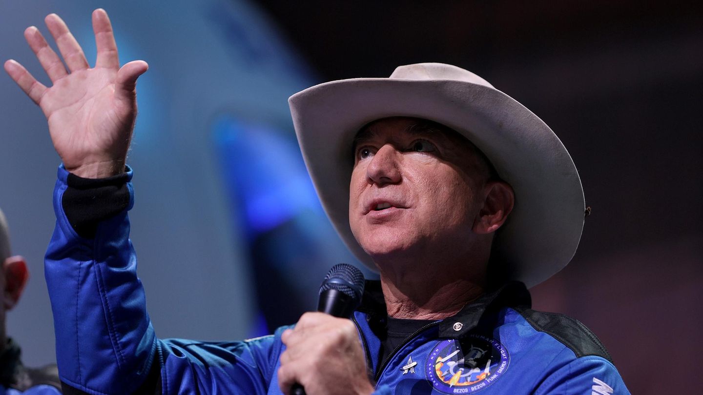 Jeff Bezos im Raumanzug und mit Cowboyhut hebt seine Hand in den Himmel