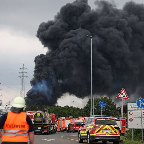 Einsatzfahrzeuge der Feuerwehr stehen unweit einer Zufahrt zum Chempark über dem eine dunkle Rauchwolke aufsteigt