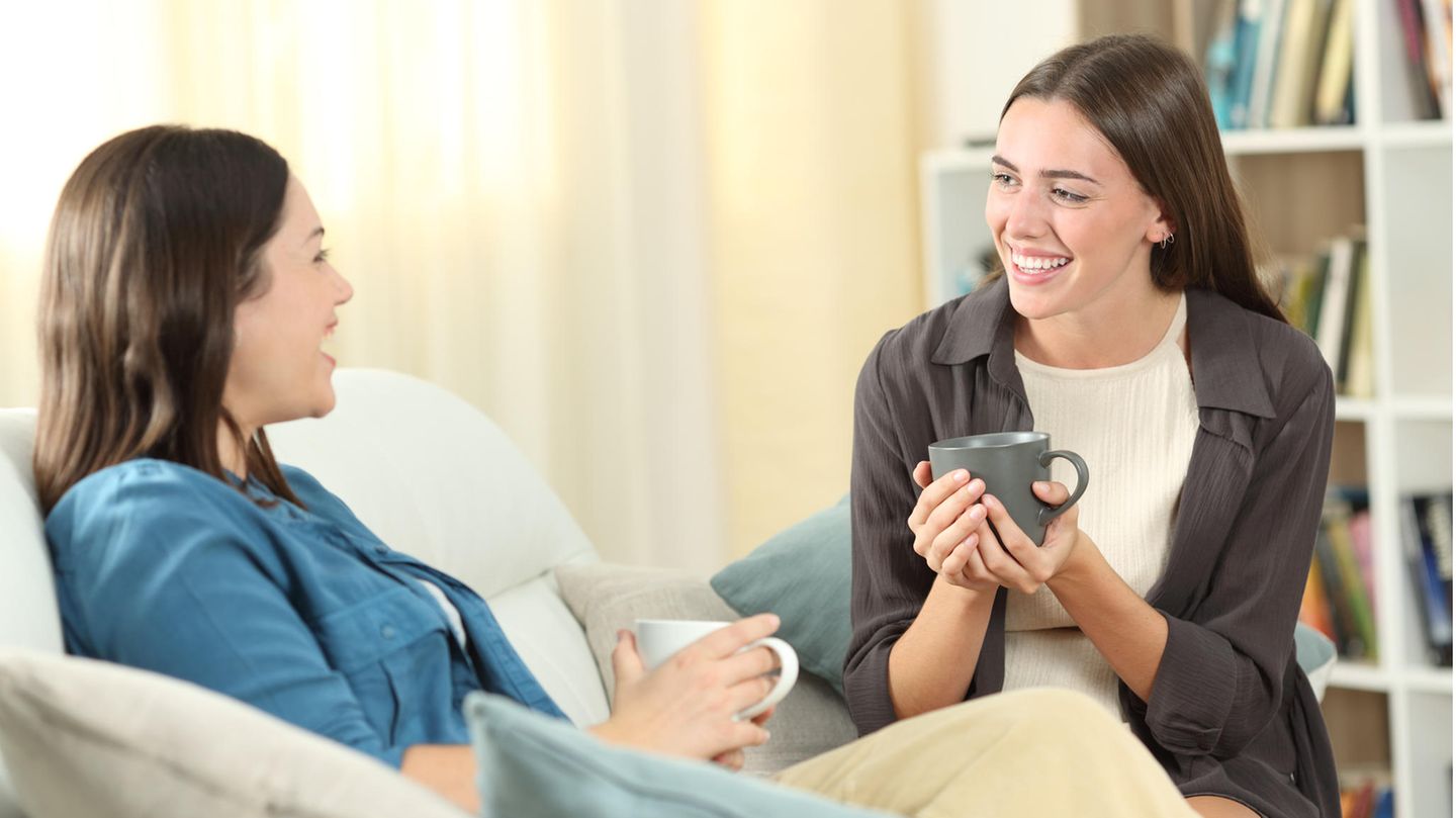 Zwei junge Frauen sitzen mit Tassen in der Hand auf dem Sofa und unterhalten sich glücklich.