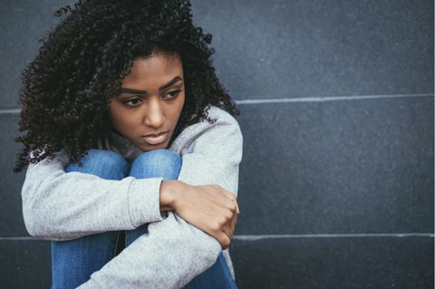 Eine junge schwarze Frau hockt an einer Wand auf dem Boden und sieht traurig aus
