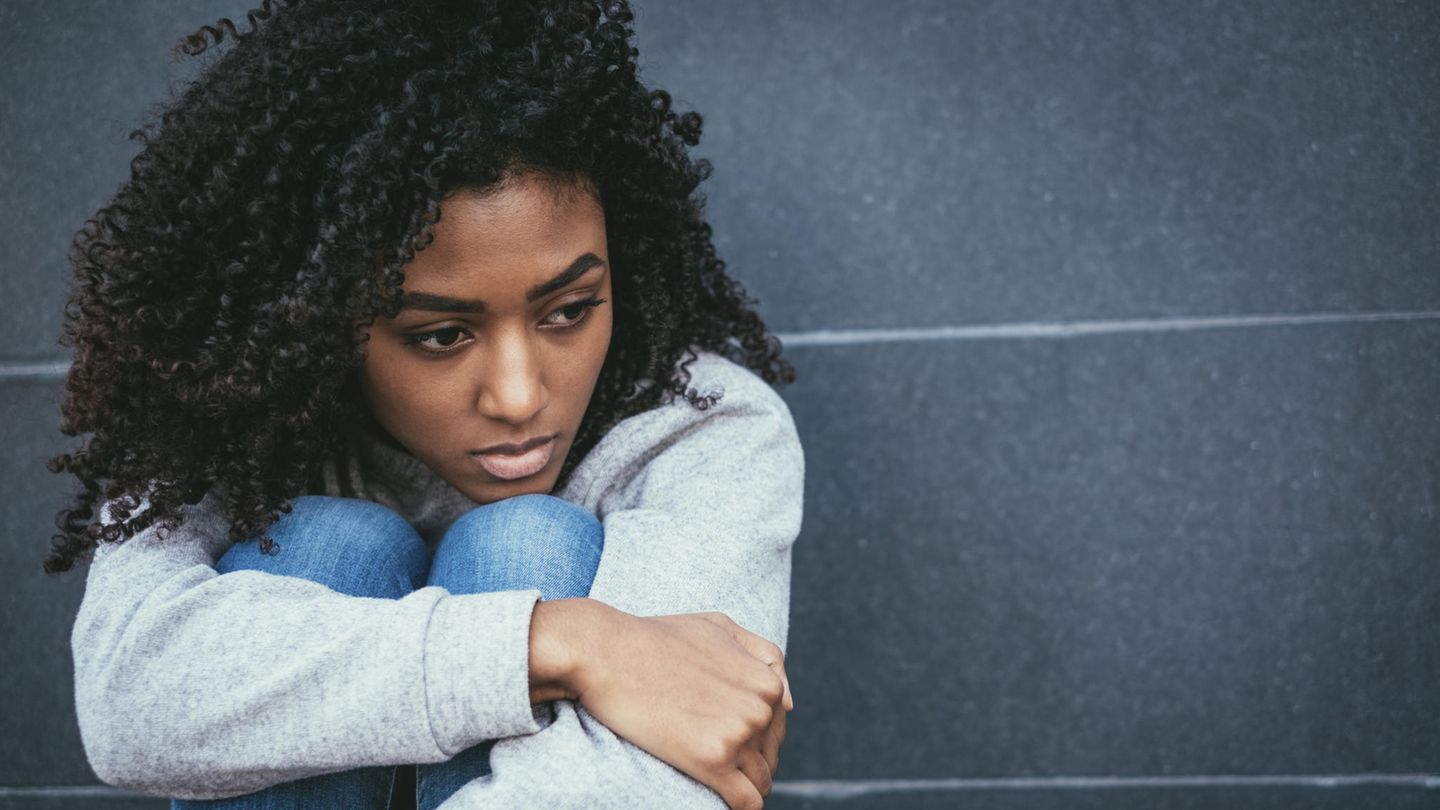 Eine junge schwarze Frau hockt an einer Wand auf dem Boden und sieht traurig aus