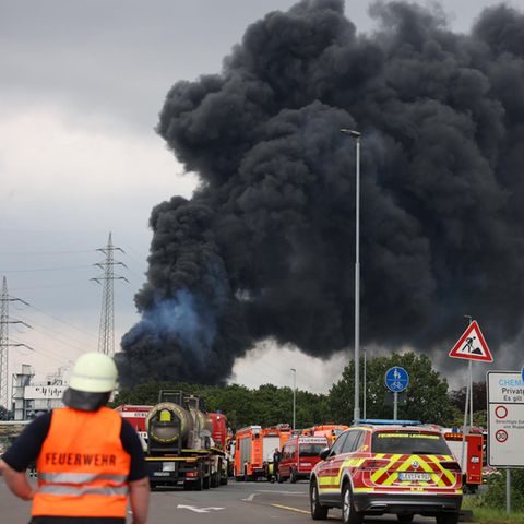 Einsatzfahrzeuge der Feuerwehr stehen unweit einer Zufahrt zum Chempark über dem eine dunkle Rauchwolke aufsteigt