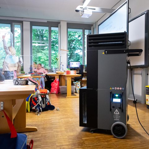 Ein schwarz-grauer Quader auf Rollen und mit Luftschlitzen im oberen Bereich steht in einem Klassenzimmer mit Grundschülern