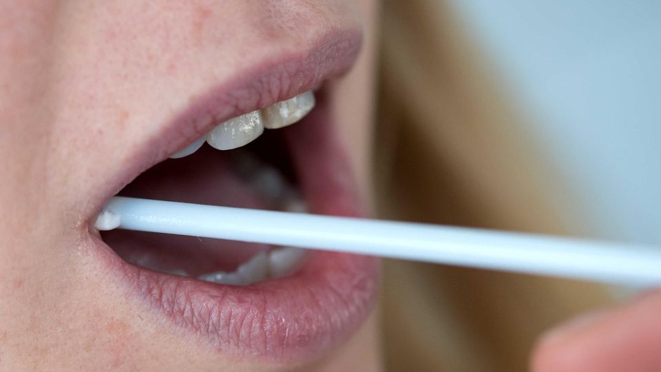 Eine Hand steckt einer blonden Frau einen Wattestab am rechten Mundwinkel in den Mund