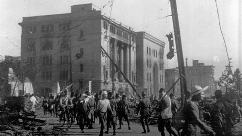 Ein Foto in Schwarzweiß zeigt Menschen, die an einem stark zerstörten Gebäude vorbeigehen