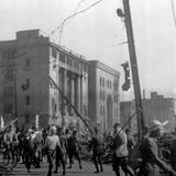 Ein Foto in Schwarzweiß zeigt Menschen, die an einem stark zerstörten Gebäude vorbeigehen