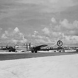 Auf dem Foto in Schwarzweiß ist ein Flugplatz mit einem Flugzeug, einem Militärfahrzeug und Menschen zu sehen