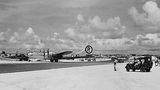 Auf dem Foto in Schwarzweiß ist ein Flugplatz mit einem Flugzeug, einem Militärfahrzeug und Menschen zu sehen
