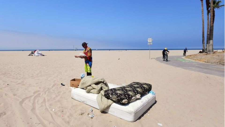 Ein Mann steht am Strand und liest, davor ist eine Matratze mit Decken zu sehen
