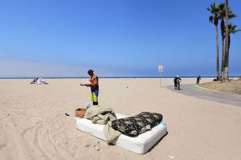 Ein Mann steht am Strand und liest, davor ist eine Matratze mit Decken zu sehen