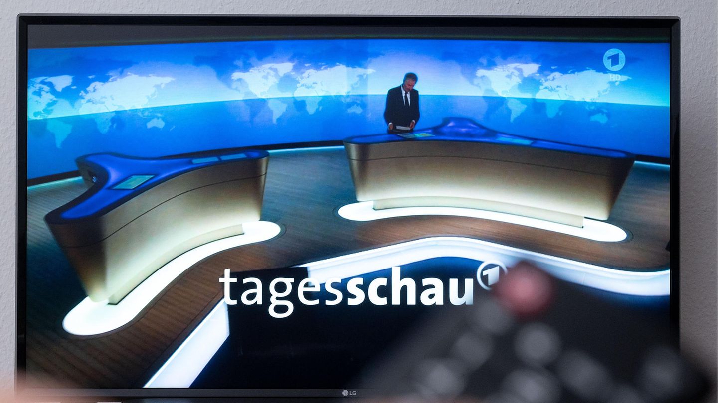 Während im Hintergrund auf einem TV-Gerät die Tagesschau beginnt, hält im Vordergrund eine linke Hand eine Fernbedienung