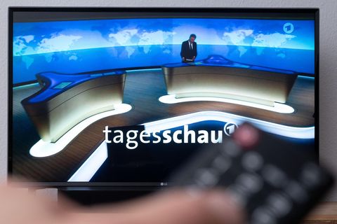 Während im Hintergrund auf einem TV-Gerät die Tagesschau beginnt, hält im Vordergrund eine linke Hand eine Fernbedienung