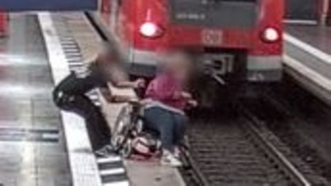 Ein Mann hält eine Rollstuhlfahrerin fest, um den Absturz ins Gleisbett zu verhindern