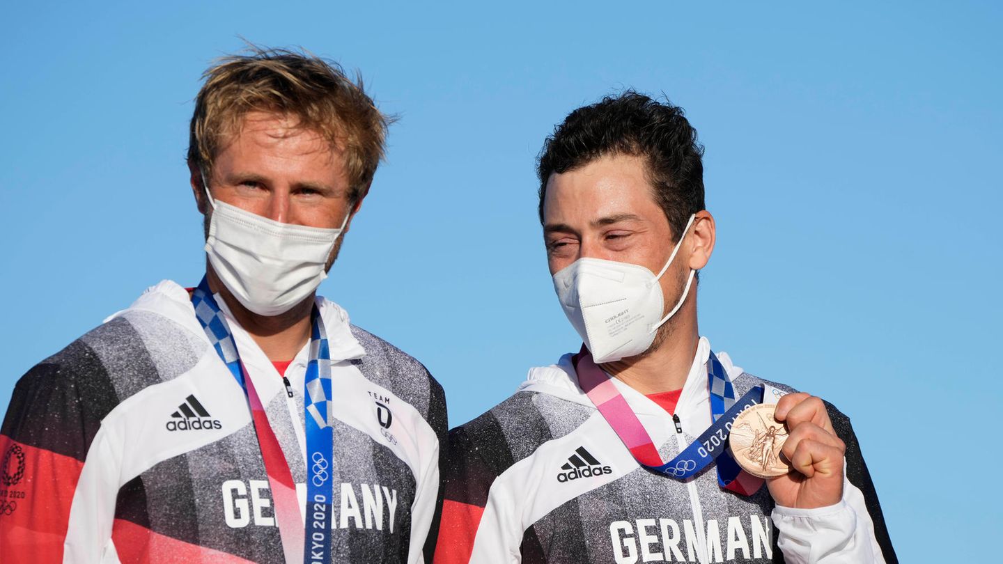 A blond white man and a dark-haired white man stand in front of a blue sky with bronze medals around their necks