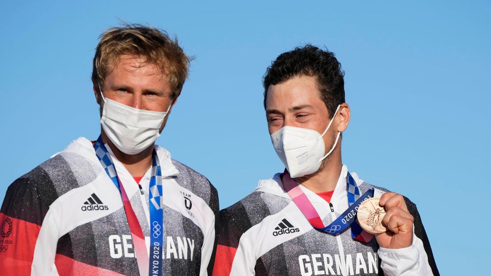Ein blonder weißer Mann und ein dunkelhaariger weißer Mann stehen mit Bronze-Medaillen um den Hals vor blauem Himmel