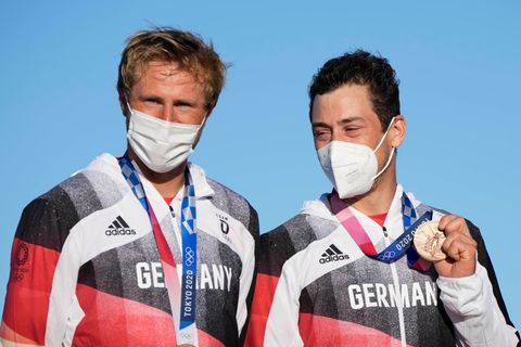 Ein blonder weißer Mann und ein dunkelhaariger weißer Mann stehen mit Bronze-Medaillen um den Hals vor blauem Himmel