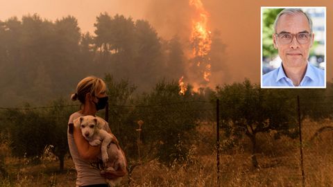 Eine blonde Frau mit schwarzem Mundschutz trägt einen weißen Hundewelpen im Arm, während im Hintergrund ein Wald brennt