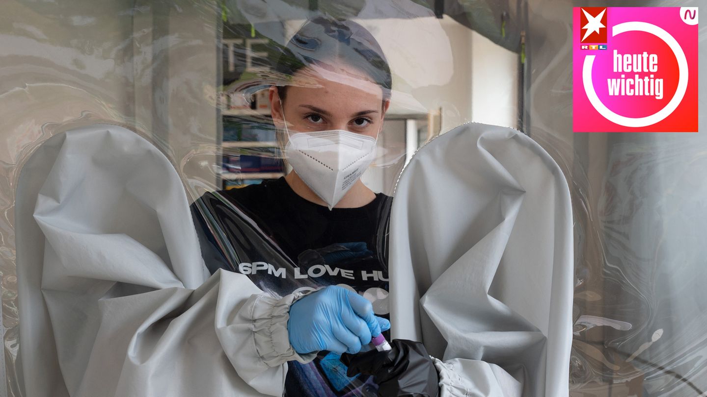 Eine junge Frau mit zurückgekämmten braunen Haaren steht mit FFP2-Maske hinter durchsichtiger Folie an einer Corona-Test-Station