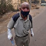 Ein älterer Mann steht mit versengter Halbglatze und weiß verbundenen Händen auf einer Straße neben verbanntem Buschwerk