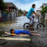 Auf einer überschwemmten Straße liegt ein schwarzhaariger Junge auf einem Surfbrett, während ein Jugendlicher Rad fährt