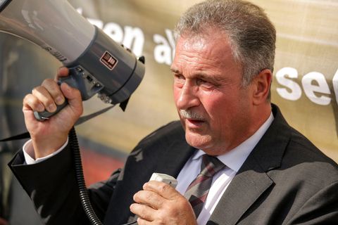 Ein weißer Mann in Anzug und Krawatte steht im Sonnenschein, während er in ein Megafon spricht. Er hat einen graue Haare