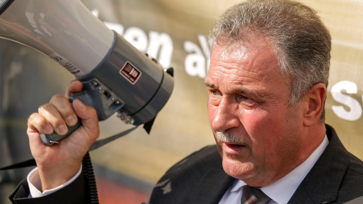 Ein weißer Mann in Anzug und Krawatte steht im Sonnenschein, während er in ein Megafon spricht. Er hat einen graue Haare