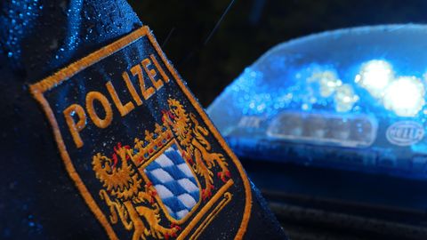 Auf einem regennassen Ärmel prangt das Wappen der Polizei Bayern, während dahinter ein Blaulicht die Dunkelheit erhellt